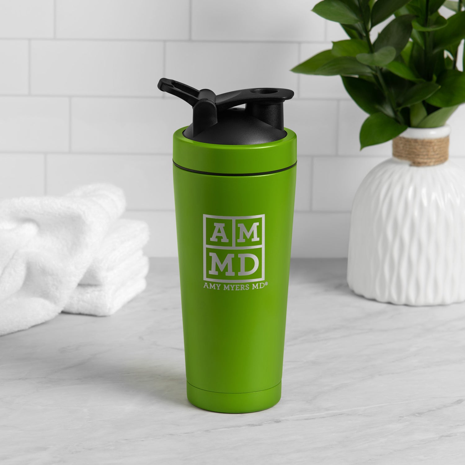 A green and silver Amy Myers MD branded shaker bottle is shown sitting on a light gray kitchen counter with a white tile backsplash.